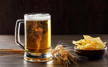 Кружка светлого пива с пеной, колосья пшеницы и миска чипсов на деревянном столе